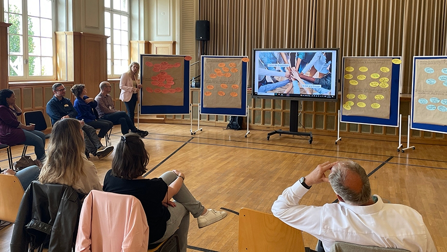 A group of adults sit in chairs in a semi-circle looking at a screen with a presentation and board with post-it notes on them