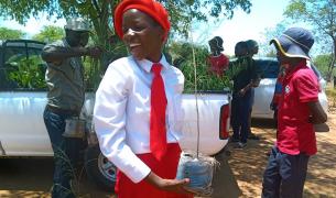 A student in school uniform carries a tree sapling in front of a pick-up with other saplings in it's bed while other students and adults gather around in the background