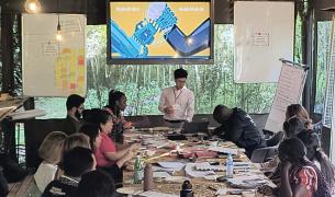 A man stands at the head of a long table with people sitting on either side, there is a screen behind him with a colorful presentation and white board and flipcharts with writing on either side