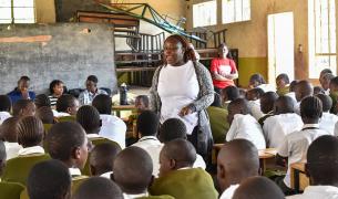 A young African woman in a white shirt and grey sweater stands in the middle of a classroom full of students