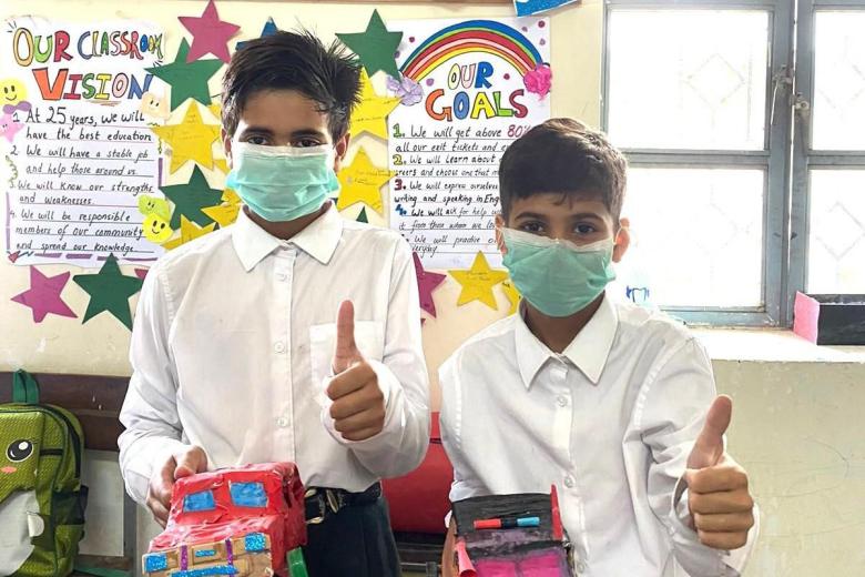 Two boys with brown skin and dark hair hold vehicles made out of cardboard and give the thumbs-up sign