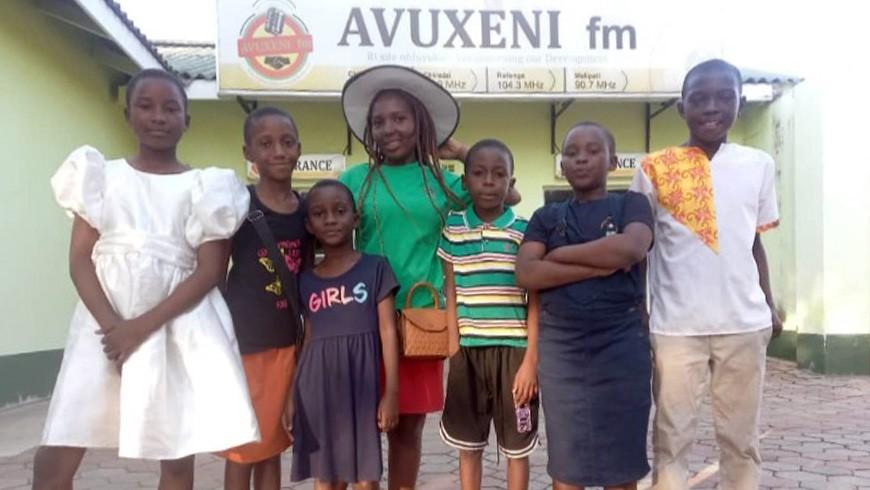 A group of students stand in front of a building with the sign AUXENI fm