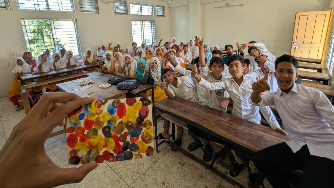 Classroom full of students smiling and posing in the background while in the foreground a hand holds up a small painting of colorful dots