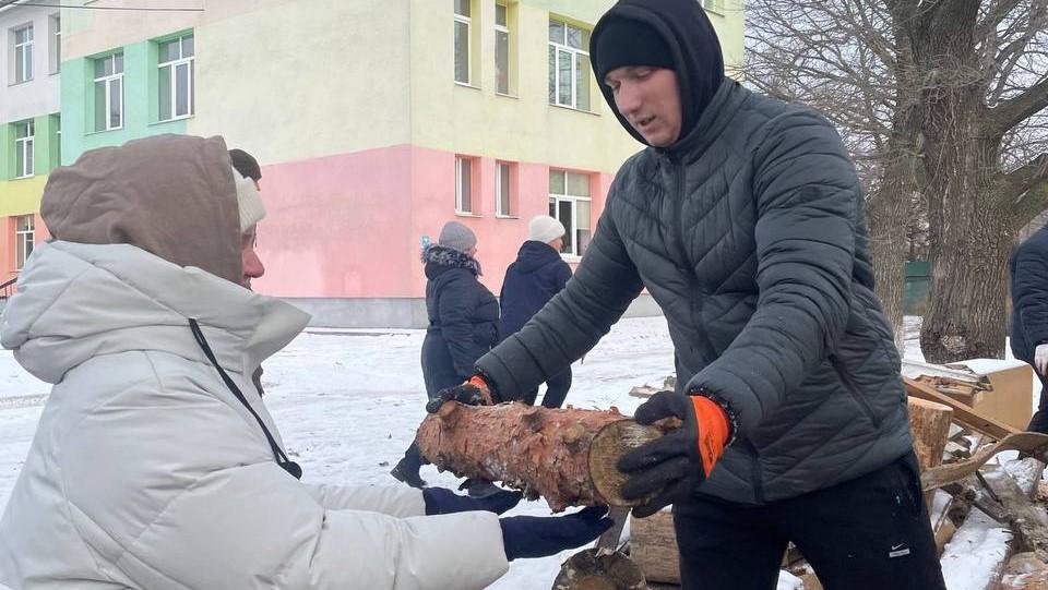 A woman in a black winter jacket and hood hands a log to another person in a light grey winter jacket and hat