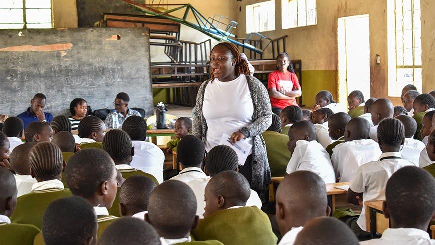A young African woman in a white shirt and grey sweater stands in the middle of a classroom full of students